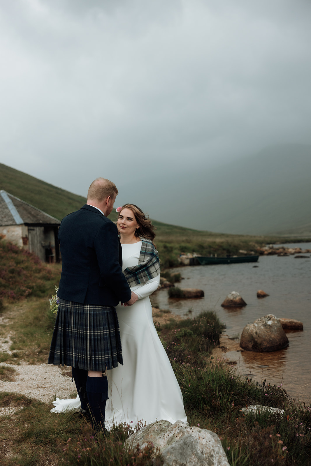 Bride and groom standing by Loch Iorsa with boathouse in background.