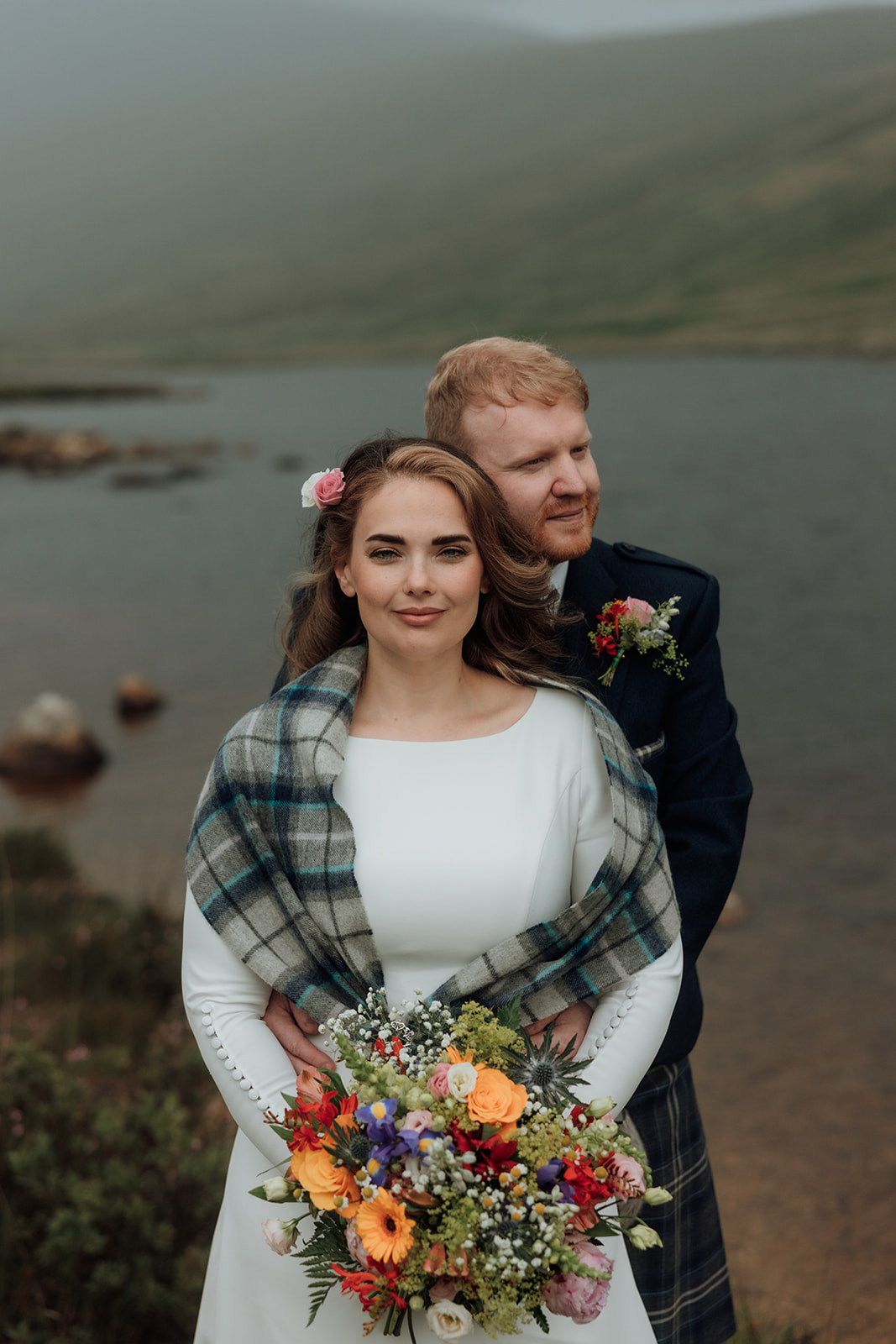 Bride holding bouquet with groom standing behind her by Loch Iorsa, Isle of Arran.