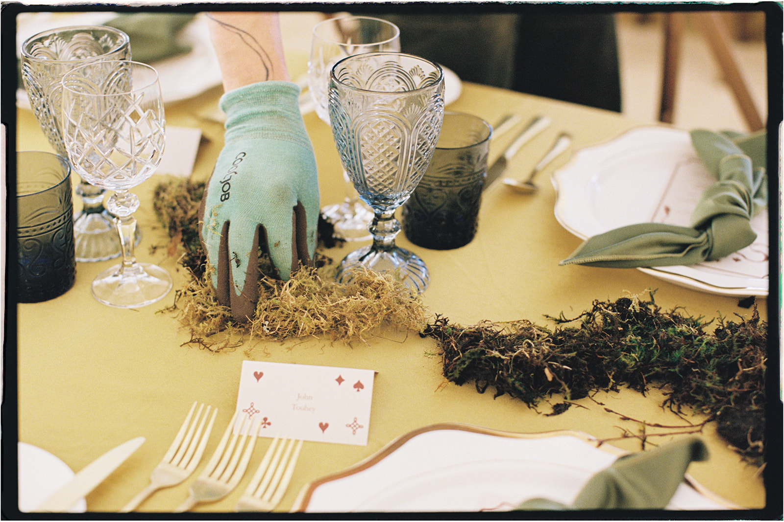 Pair of gardening gloves resting on moss beside a yellow-linen table setting, photographed on 35mm film.