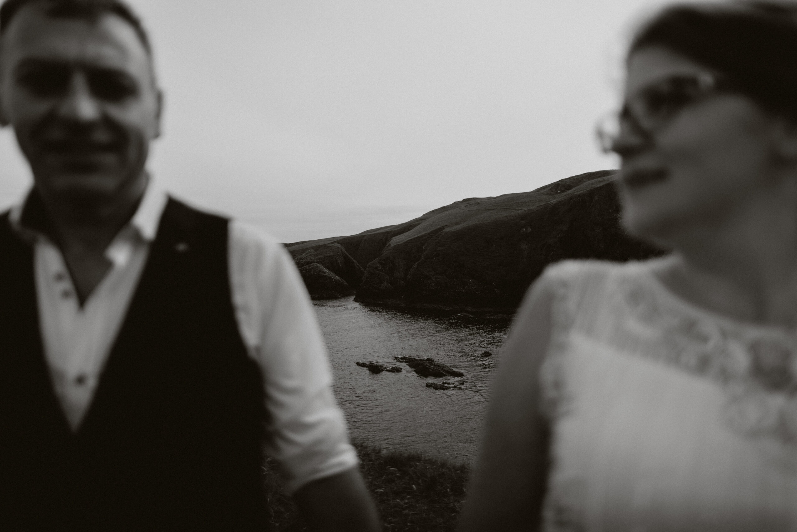 wedding couple stand out of focus in the foreground with coastline in focus behind them